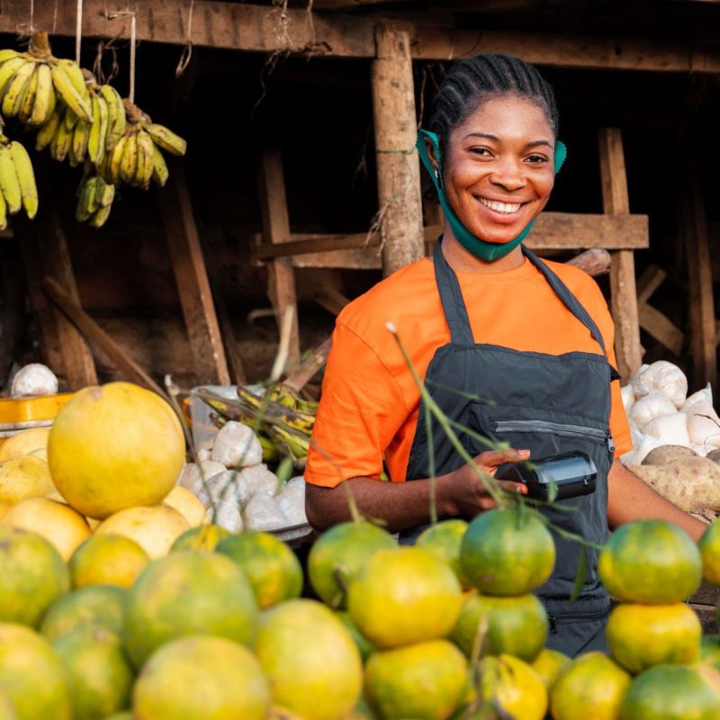 Une commerçante souriante devant son étal d’agrumes et de bananes